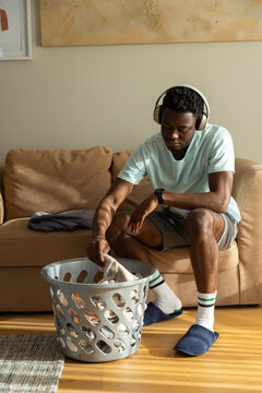 African-American man 20s leaning and picking clothes from grey laundry basket on living room floor