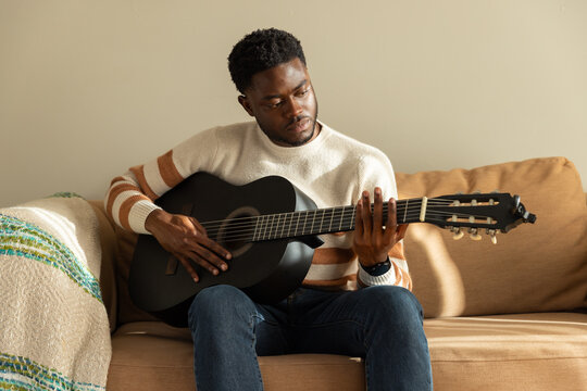 African man sitting on sofa in living room wearing cream sweater strumming matte black guitar