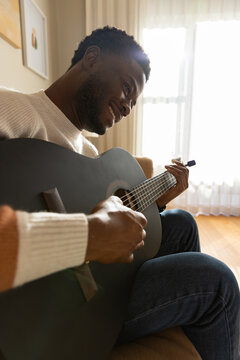 Man wearing cream knit sweater and jeans, playing dark acoustic guitar with capo on couch