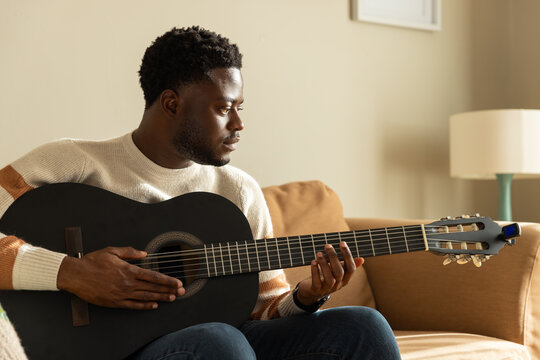 African American man playing black guitar on tan sofa wearing cream sweater with clip-on tuner