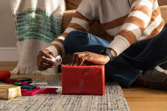 African American man in striped sweater, jeans sitting on rug wrapping red gift with tape dispenser