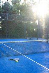 Obraz premium Black padel racket and two yellow balls are resting on blue court near net, sun glare