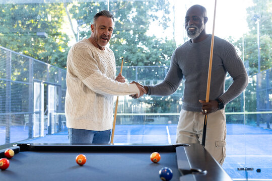 Diverse male friends shaking hands at pool table, holding cues, billiard balls on felt