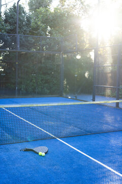 Black padel racket and two yellow balls are resting on blue court near net, sun glare