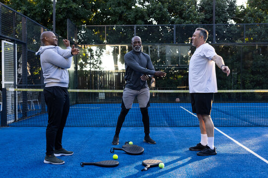 Diverse male teammates stretching, warming in gear on paddle court near low net with paddles, balls
