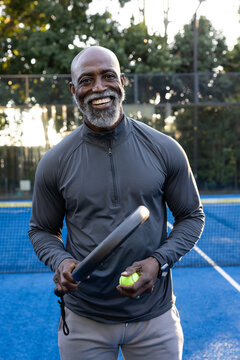 Mature African man smiling, holding solid paddle and yellow ball on blue court wearing zip top