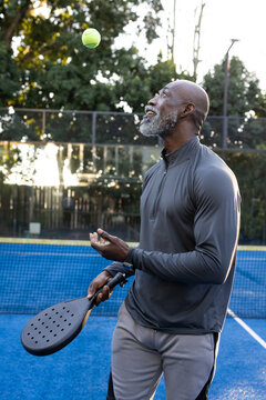 Mature man tossing neon yellow ball while holding black paddle, wearing watch on blue court