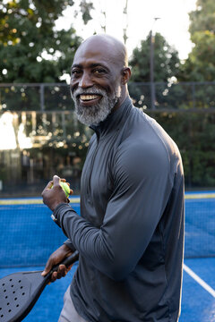 Senior male wearing zip-up top standing on blue pickleball court holding paddle and yellow ball