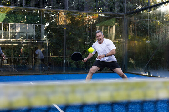 Adult male lunging swinging in white shirt on blue padel court holding paddle hitting yellow ball