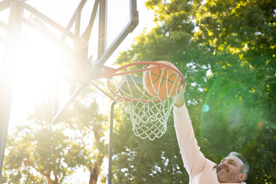 Adult man reaching and pushing basketball through rim at outdoor park court, lens flare, copy space