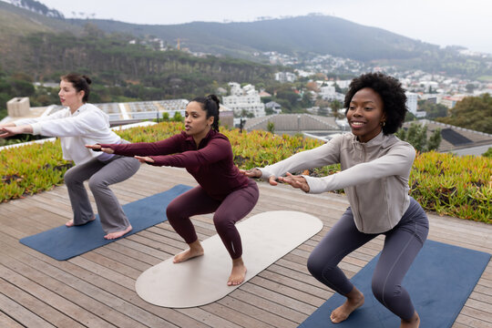 Diverse female friends wearing activewear doing chair pose on roof deck on navy beige blue mats