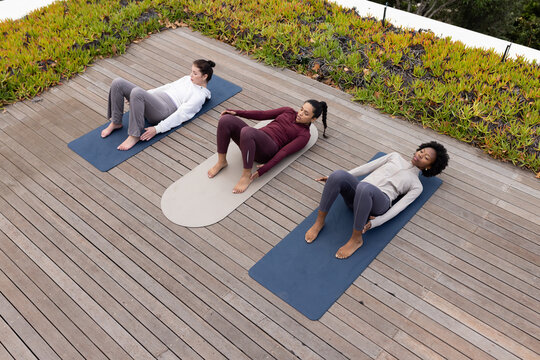Three adult women wearing athletic clothes lying on mats engaging core on wood deck smartphone near