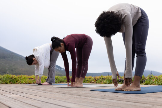 Female friends in activewear practicing yoga on wooden deck by coastal hills with mats, copy space