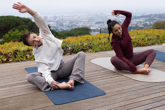 Diverse female friends stretching on navy gray mats on wood deck in athletic wear overlooking water