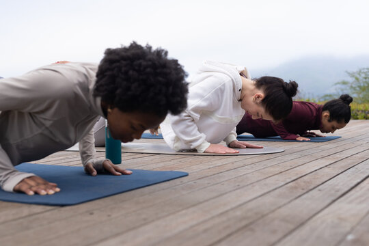 Diverse female friends practicing low push-ups on dark blue mats on wooden deck with water bottle
