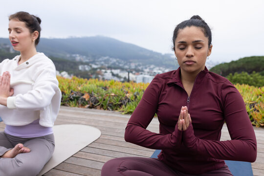 Diverse women meditating on deck overlooking town on mats, maroon zip, white hoodie, copy space