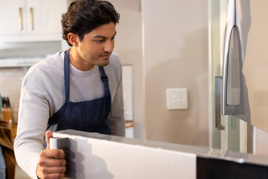 Adult African man in dark denim apron pulling oven door, peering inside in kitchen near fridge