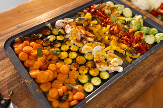 Metal baking tray is holding seasoned sliced vegetables on wooden countertop, knife handle visible