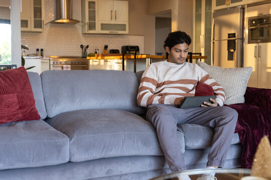 African man sitting on right side of gray sofa holding tablet wearing striped sweater, copy space