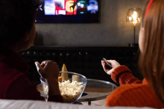 Diverse female friends in knit sweaters on couch at home holding remote, watching TV