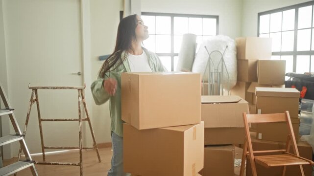 Woman holds stacked moving boxes inside a sunlit building room with ladder, folding chair and packed cartons; new beginning hopeful.