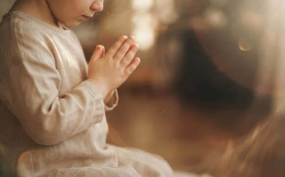 Young Caucasian child in linen clothing praying with hands clasped together in soft warm indoor light