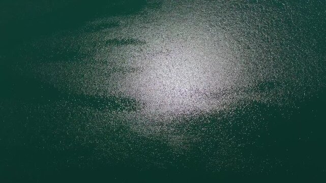 Top-down drone clip of glowing sunrays reflecting off a shimmering turquoise water surface, creating abstract aquatic background with wind ripples