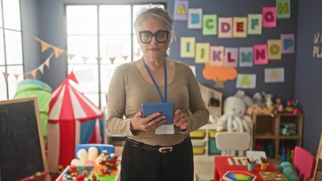 Woman teacher with short gray hair and black glasses holding tablet with both hands in classroom full of toys, tent and alphabet display; calm caregiving.