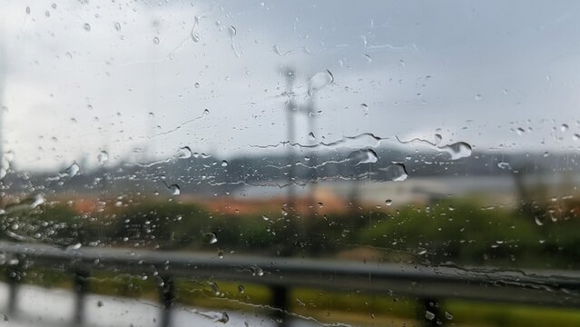 Abstract close-up of water droplets and rain streaks on a vehicle window with blurred green hills and rural farmland in background.