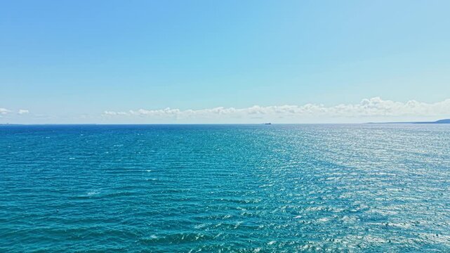 The ocean is calm and blue with gentle waves. A ship can be seen far away on the water. The sky is clear with some clouds. It is a sunny day with no signs of rain.