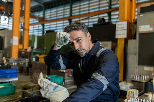 Tired industrial worker resting during shift in manufacturing factory, showing fatigue, workplace stress, physical labor, occupational health, safety and demanding working conditions.