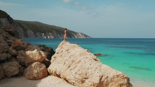 Beautiful woman standing on a giant rock above Kefalonia bay, admiring turquoise sea and cliffs in Greece