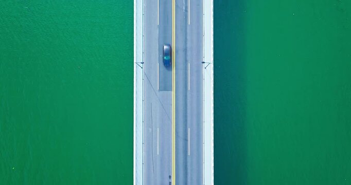 Top-down aerial view of an empty gray bridge with yellow centerline crossing calm turquoise water in bright sunlight.