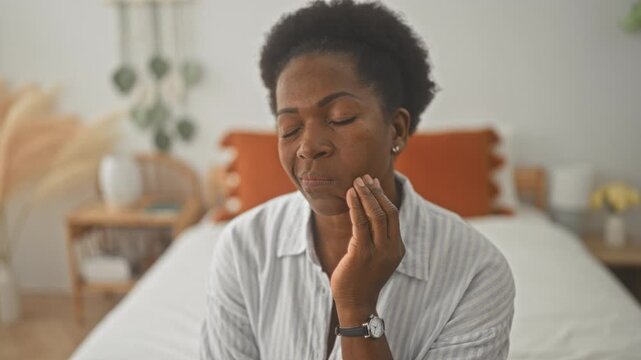 Woman touching cheek and wincing on a bed, hand on jaw and mouth open for toothache in building; pain concern.