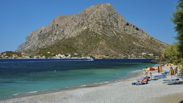 Massouri, umbrellas and sun beds on a pebble beach with a distinctive rock in the background, Kalymnos, Sponge Diving Island, Dodecanese, Greek Islands, Greece