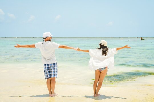 Caucasian couple man woman stand together arms outstretched enjoy white sand beach in South Zanzibar, Tanzania. Romantic summer holiday by turquoise ocean with clear blue sky. Honeymoon paradise vibes