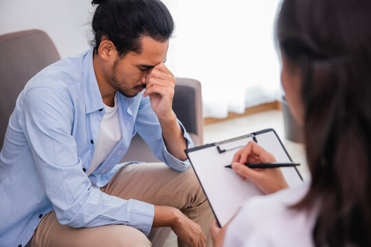 Female psychologist taking notes while counseling a stressed Asian man during therapy session