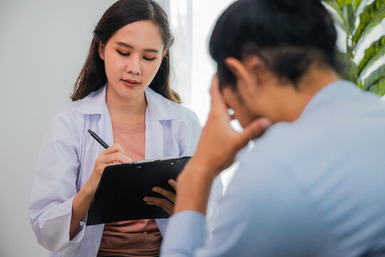 Female psychologist taking notes while counseling a stressed Asian man during therapy session
