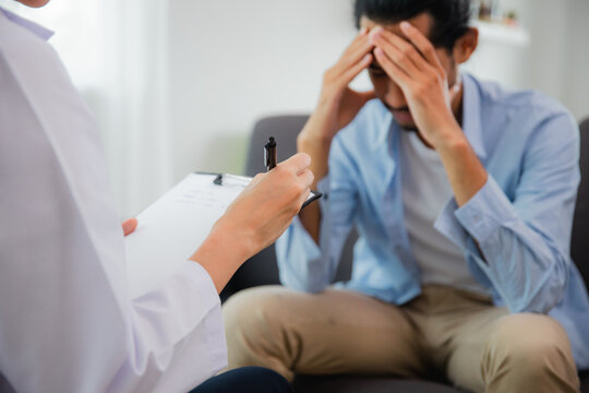 Female psychologist taking notes while counseling a stressed Asian man during therapy session