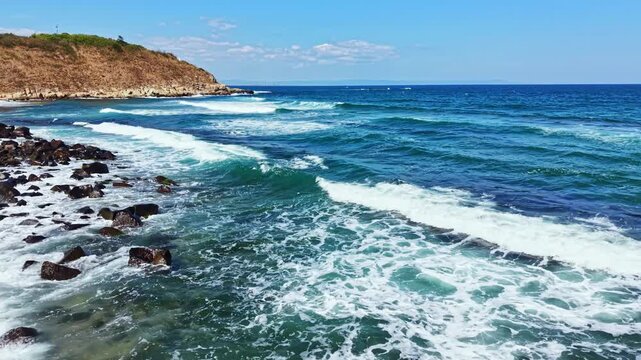 Water crashes against rocks as waves come ashore. The sun shines down on the ocean. A few clouds dot the bright blue sky.