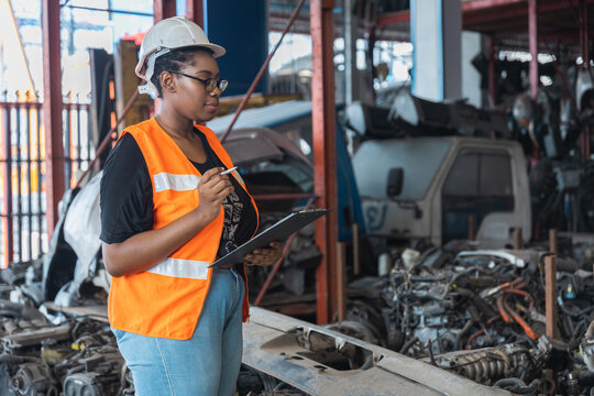 Black female inspector in safety vest checking car parts with clipboard in industrial warehouse