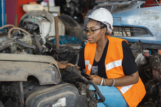 Black female inspector in safety vest checking car parts with clipboard in industrial warehouse