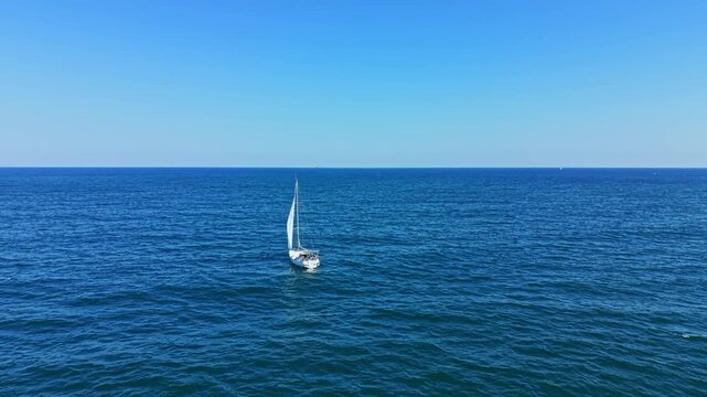 A sailboat moves across the ocean with no other boats in sight. The sky is clear and blue, and the water is deep blue. The sun shines down, creating a bright scene during the day.