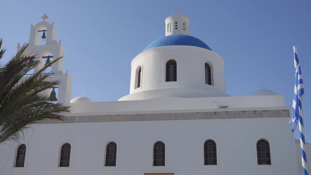 Stunning white greek church with blue dome and traditional bell tower under clear sky, greek flag waving next to it, santorini, greece.
