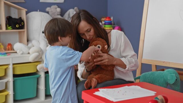 Woman holding plush monkey smiles and interacts with a little boy in a preschool classroom with easel and toys; nurturing learning.