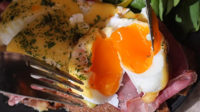 Delicious eggs benedict with runny yolk being cut by fork and knife on toast with meat
