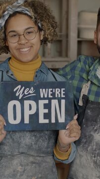 Man and woman in dirty aprons holding Yes we are open banner smiling looking at camera in pottery workshop. Creative work and business concept.