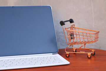 Miniature shopping cart next to a modern tablet with blank mockup screen on a wooden table, online shopping and e-commerce business concept with copy space. © Isman