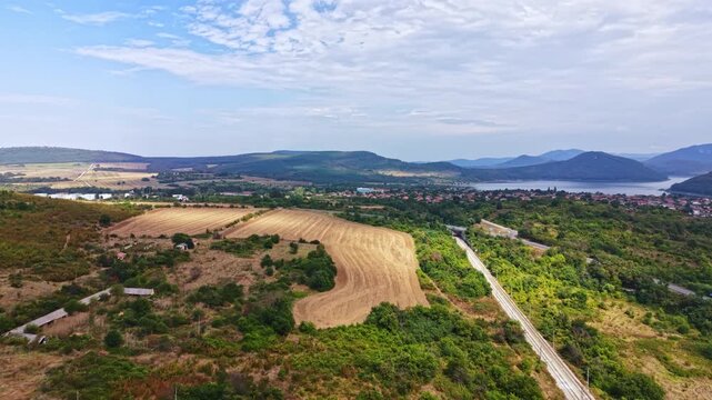 The landscape shows open fields and hills with a distant water body. A train track runs through the area. The sky has clouds, indicating a clear day.