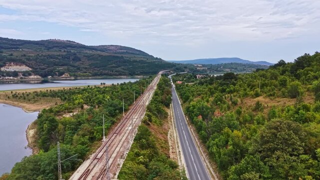 The railway tracks run parallel to a road beside a river surrounded by hills and trees. The sky shows signs of clouds suggesting a pleasant day for travel.